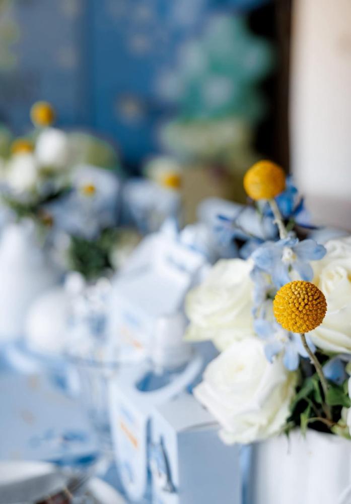 Detail of blue and white christening table styling with white roses, craspedia, favour boxes and glassware