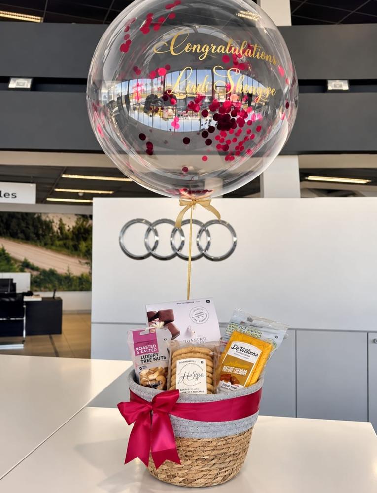 Personalised congratulations confetti bubble balloon above a gourmet gift basket delivered to a motor dealership showroom
