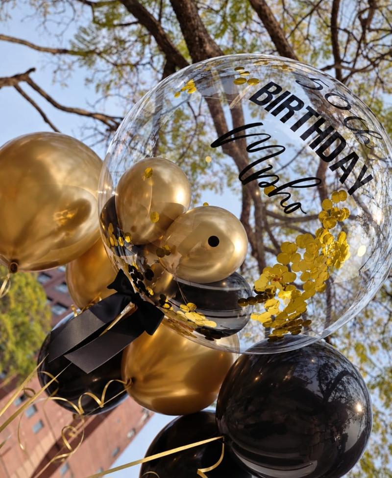 Personalised gold-lettered clear confetti bubble balloon in a gold and black balloon bouquet photographed outdoors