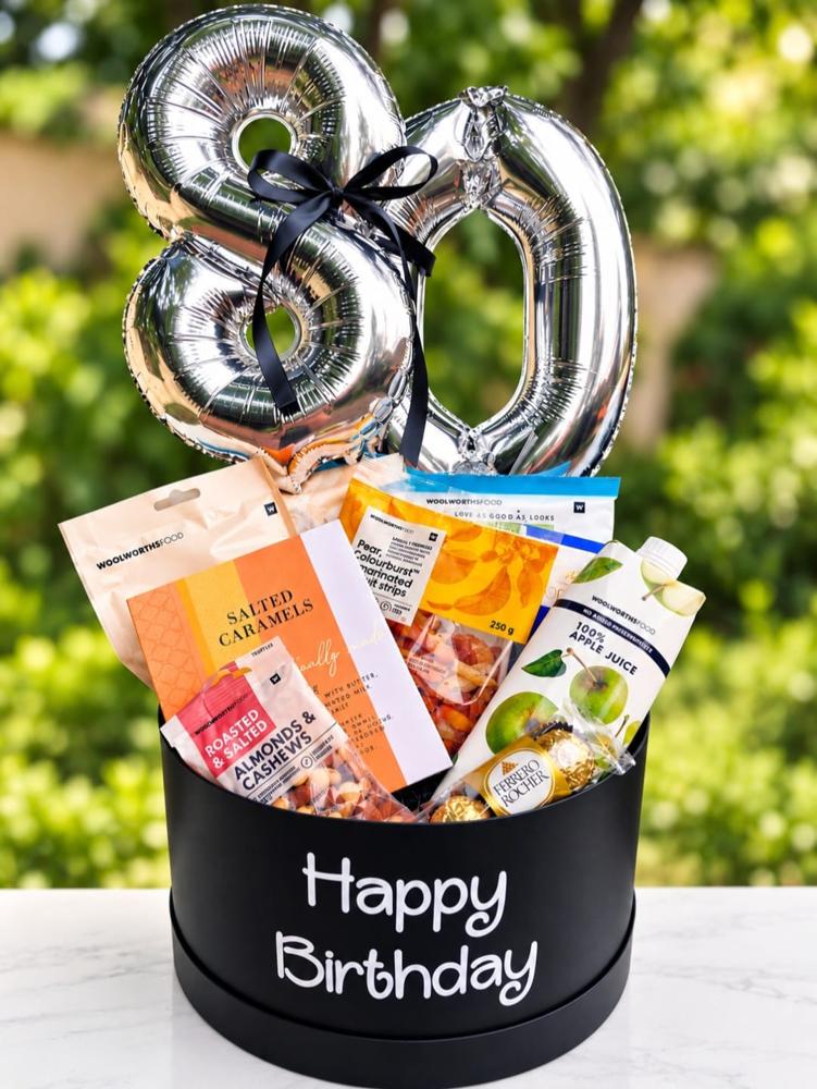 Silver 80 number balloons tied with black ribbon above a black Happy Birthday hat box filled with gourmet treats and juice