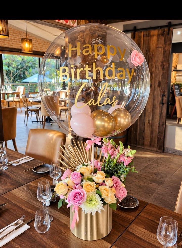 Personalised gold-lettered bubble balloon above a pastel rose and chrysanthemum arrangement on a set restaurant table