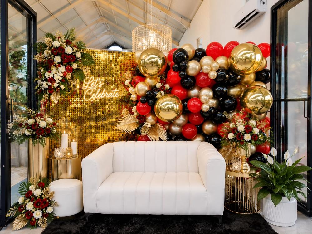Red, black and gold balloon wall beside a gold sequin backdrop and white lounge sofa at an event
